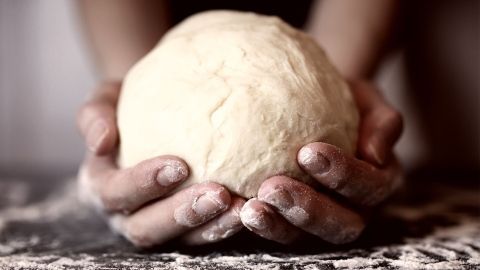 Ein gut geformter Pizzateigball wird von Händen gehalten, umgeben von einer bemehlten Arbeitsfläche.