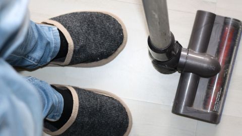 In gemütlichen Schlosspantoffeln steht eine Person neben einem Staubsauger auf einem Laminatboden und bereitet sich zum Saubermachen vor.