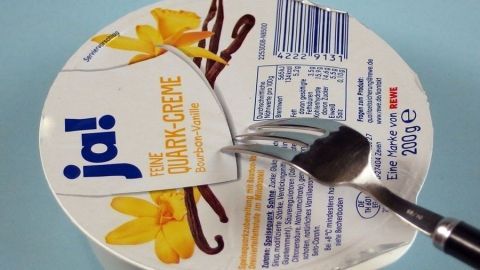 Ein Joghurtbecher wird mit einer Gabel geöffnet, dabei spritzt nichts, auf einem hellblauen Hintergrund.