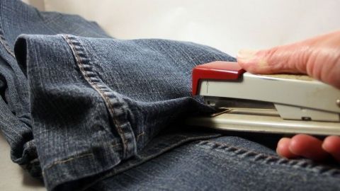 Eine Hand kürzt eine zu lange Hose mit einem Hefter, während sie auf einer neutralen Oberfläche liegt.