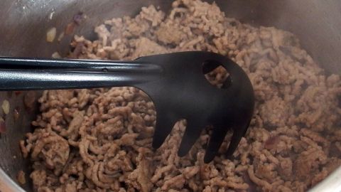 Hackfleisch wird in einem Topf krümelig angebraten, während ein schwarzer Kochlöffel das Fleisch umrührt.