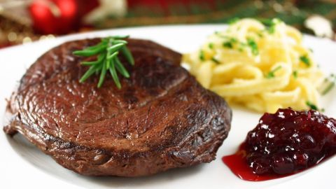 Ein saftiger Hirschbraten liegt auf einem Teller, garniert mit Rosmarin, neben Nudeln und Preiselbeermarmelade.