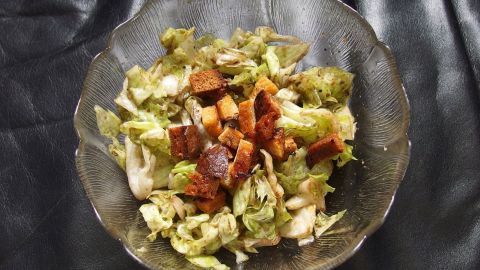 Ein Salat mit Stücken von altbackenen Brötchen wird in einer Glas-Schüssel serviert. Der Salat liegt auf einem schwarzen Untergrund.