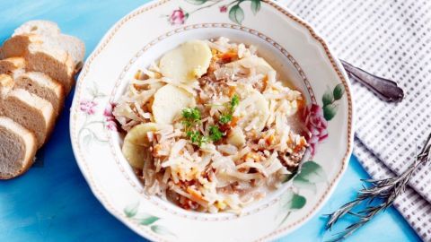 Eine Sch&uuml;ssel Wei&szlig;kohlsuppe mit Fleisch und Kr&auml;utern steht auf einem blauen Tisch, umgeben von Brot und einer Serviette.