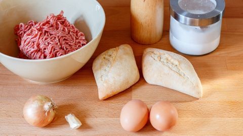 Gemischtes Hackfleisch liegt in einer Schüssel, während Zwiebel, Brötchen, Eier und Gewürze zur Zubereitung bereitstehen.