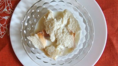 Eine Schale mit süßer, cremiger Milch und kleinen Stückchen, auf einem weißen Teller, liegt auf einem orangen Tisch.