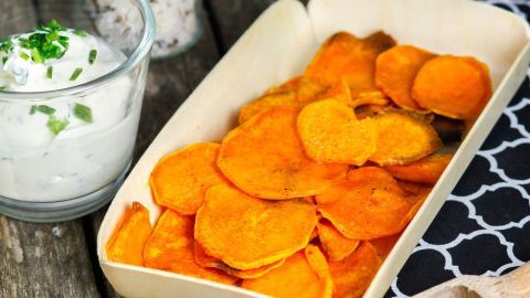 Süßkartoffelchips liegen in einer klobigen Schale und werden neben einer Glas-Schale mit Dip auf einem Holzuntergrund präsentiert.