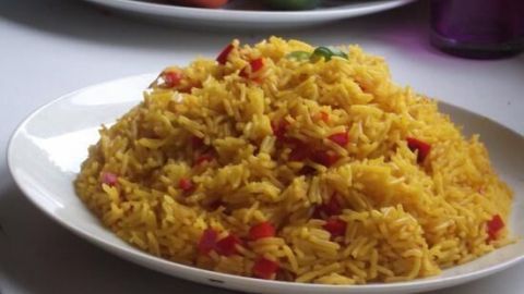 Ein gelb gefärbter Reis mit roten Paprikastückchen wird appetitlich auf einem weißen Teller präsentiert, umgeben von frischem Obst.