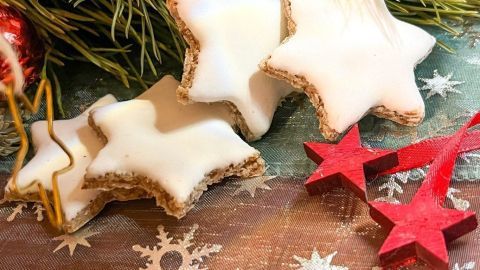 Zimtsterne liegen festlich dekoriert inmitten von grünen Tannenzweigen und roten Weihnachtssternen auf einem winterlichen Tisch.