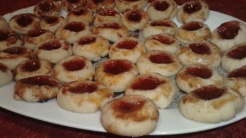 Kleine, runde Plätzchen mit roter Marmelade in der Mitte liegen auf einem weißen Teller, vor einem roten Tisch.