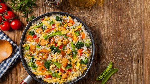 Eine große Pfanne enthält bunte Zutaten wie Reis, Gemüse und Hähnchen, um eine süß-saure China-Pfanne zuzubereiten. Tomaten und Öl ergänzen die Szene.