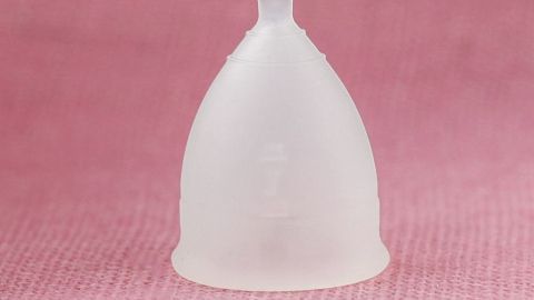 In 10 Jahren braucht eine Frau 3.500 Tampons oder Binden oder eine Menstruationstasse! Die Tasse kostet nur einmalig 13 - 35 Euro und spart somit bares Geld. Eine Menstruationstasse steht auf einem rosa Hintergrund, symbolisiert kostengünstige Menstruationspflege und Nachhaltigkeit.