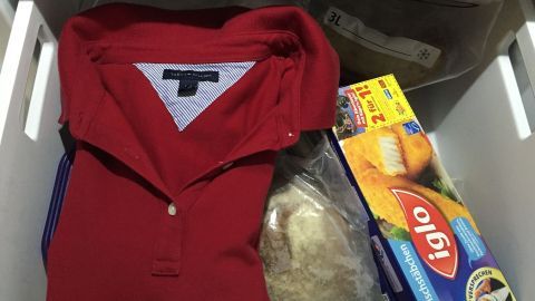 T-Shirt für einen Tag in den Gefrierschrank stecken und am nächsten Tag mit der Handfläche glatt streichen. Ein rotes Polo-Shirt liegt zusammen mit gefrorenen Lebensmitteln und einer Verpackung in einer Tiefkühltruhe.