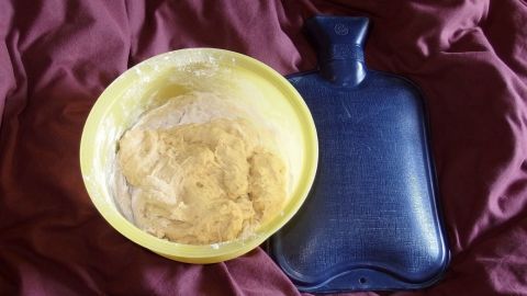 Ein gelber Schüssel mit Hefeteig liegt auf einem roten Bett, neben einer blauen Wärmflasche, die Wärme spendet.