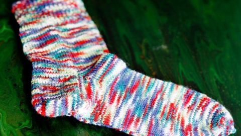 Bunte Socke liegt auf einer grünen Oberfläche und zeigt das Ergebnis des parallelen Sockenstrickens mit zwei Nadelspielen.