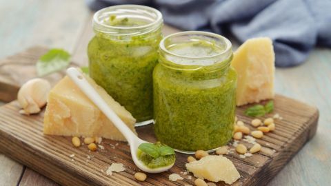 Zwei Gläser Pesto Verde stehen auf einem Holzbrett, umgeben von Parmesan und Pinienkernen, mit einem Löffel daneben.