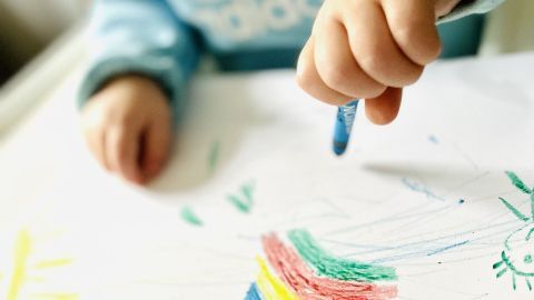Ein Kind in einem blauen Sweatshirt zeichnet mit einem Buntstift auf einem weißen Blatt Papier, umgeben von Kunstwerken.