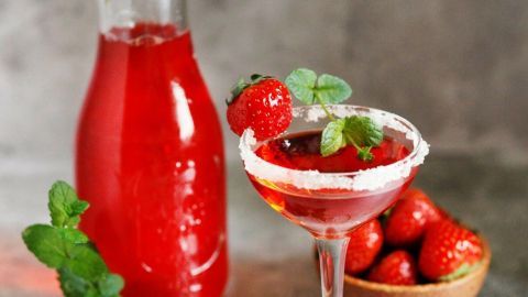 Ein elegantes Cocktailglas mit rotem Getränk und frischen Erdbeeren steht neben einer Karaffe auf einem hellen Hintergrund.