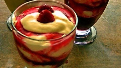 Gläser mit Schichten aus Mascarpone-Creme und rotem Obstpüree, gekrönt mit einer Himbeere, auf Holztisch.