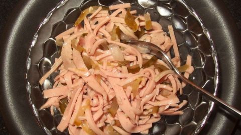 Zum Annmachen eines Wurstsalats eignet sich auch hervorragend die Gurkenbrühe aus dem Gurkenglas. Wurstsalat liegt appetitlich in einer Schüssel, während ein Löffel die Mischung aus Wurst und Gurken umrührt.