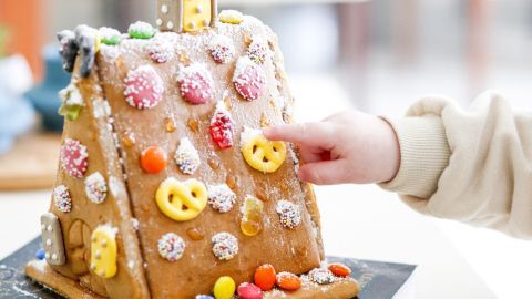 Ein kleines Kind berührt eine bunt dekorierte Lebkuchenhaus-Dekoration mit vielen Bonbons in einer hellen Umgebung.