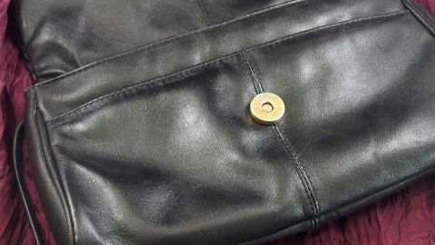Eine schwarze Ledertasche liegt flach auf einem weichen, lila Untergrund, bereit zum Färben oder Verschönern.