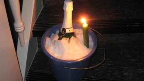 Sektflasche kühlt im Schnee-Eimer auf einer Treppenstufe, daneben brennt eine Kerze, die für stimmungsvolles Licht sorgt.