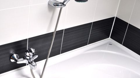 Eine glänzende Duscharmatur steht über einer weißen Badewanne, umgeben von grauen und weißen Fliesen, möglicherweise mit Kalk.