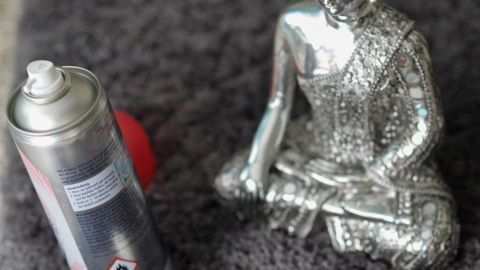 Eine silberne Buddhafigur sitzt auf einem Teppich neben einer Dose Backofenspray, während ein Schaumfleck sichtbar ist.