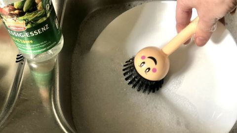 Eine Hand hält eine bunte Spülbürste, die Geschirr abwäscht, während Essig-Essenz im Spülbecken steht.