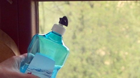 Ein Hand hält eine Flasche mit Klarspüler, während ein Fenster geöffnet ist, um den Ausblick auf die Natur zu ermöglichen.