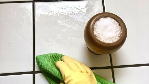 Ein gelbes Gummihandschuh hält ein grünes Tuch, während es Fliesen mit Wasser und Salz reinigt. Ein Gefäß mit Salz steht daneben.
