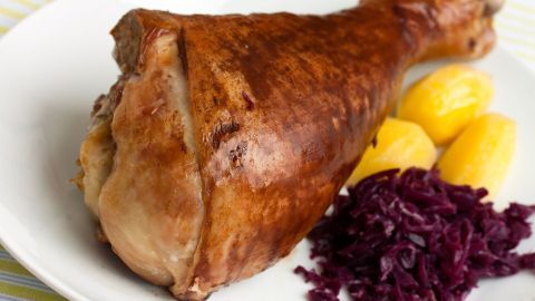 Ein gebratener Gansbeinschen liegt auf einem Teller, umgeben von Rotkohl und Kartoffeln, auf einem gestreiften Tisch.