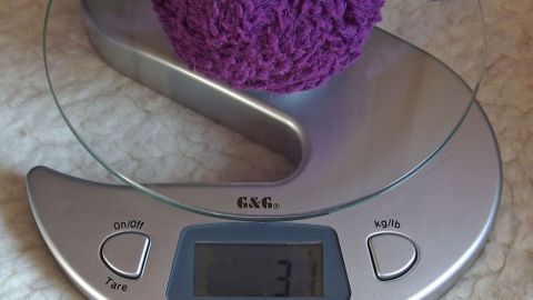Ein lila Wollknäuel liegt auf einer digitalen Waage, die 37 Gramm anzeigt, auf einem hellen, weichen Untergrund.