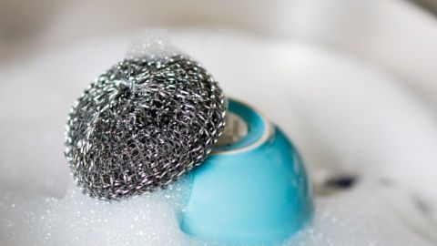 Ein blauer Topf liegt in seifigem Wasser, während ein Stahlschwamm darüber schwebt und Etiketten entfernt.