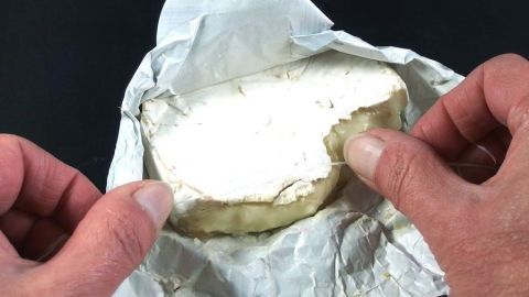Zwei Hände halten ein Stück Käse, während es aus einer Papierverpackung genommen wird, bereit für die Verwendung mit Zahnseide.