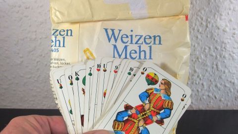 Spielkarten liegen in einer Hand, während eine Packung Weizenmehl im Hintergrund sichtbar ist.