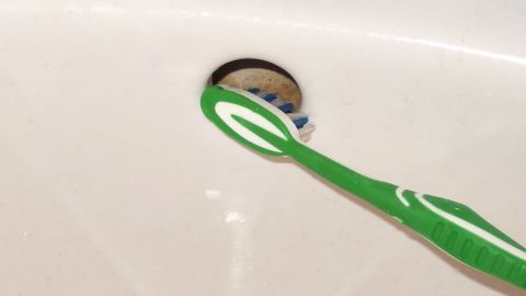 Ein grünes Zahnputzgerät wird verwendet, um den Waschbeckenüberlauf zu reinigen, während sich das Waschbecken in einem Badezimmer befindet.