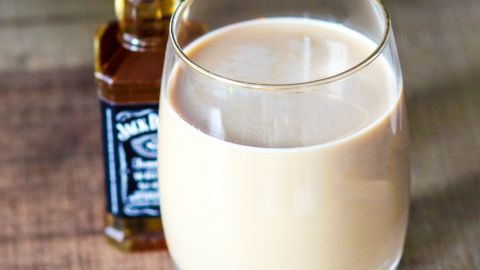 Ein transparenter Becher mit einem Getränk in cremiger Farbe steht auf einem Holztisch, neben einer kleinen Flasche mit Whiskey.