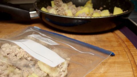 Eine gefüllte Plastiktüte mit eingefrorenem Hackfleisch und Kartoffeln liegt auf einem Holzbrett neben einer Pfanne, die brät.