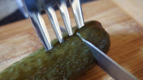 Ein Gurke wird mit einem Messer auf einem Holzbrett geschnitten, während eine Gabel sie fixiert.