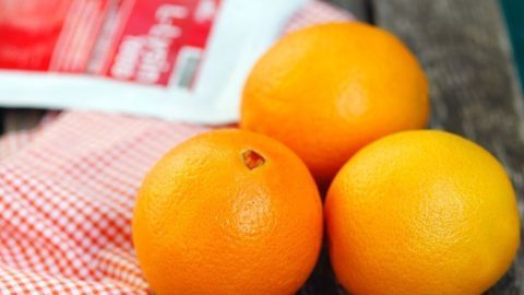 Drei saftige Orangen liegen auf einem Holztisch, während im Hintergrund ein Paket Lysin sichtbar ist.