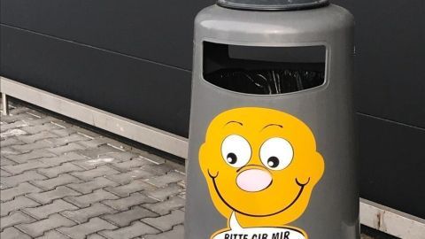Ein grau lackierter Müllbehälter steht auf einem gepflasterten Boden. Er ermutigt zum Müllvermeiden mit einem freundlichen Smiley und der Aufschrift: "BITTE GIB MIR DEINEN ABFALL".