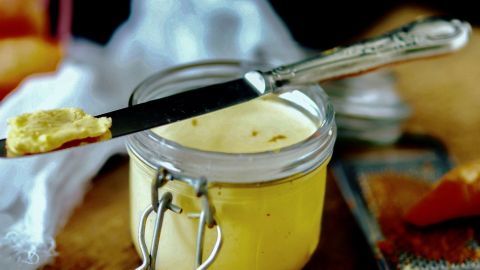 Gelbe Möhrenbutter befindet sich in einem Glas mit einem Messer, umgeben von einer Holzunterlage und anderen Zutaten.