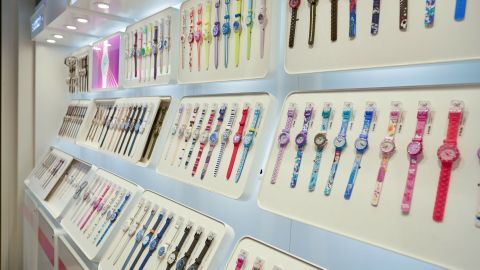 Drei Swatch Uhren sind auf einem hellen, modernen Regal ausgestellt, umgeben von weiteren bunten Uhren.