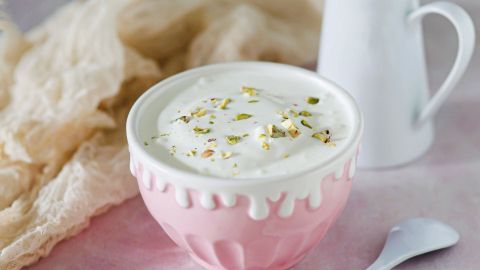 Eine rosa Schüssel mit Quarkspeise, garniert mit Pistazien, steht auf einem sanften, pastellfarbenen Hintergrund neben einer kleinen Milchkanne.