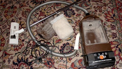 Ein defekter Staubsauger liegt auf einem Teppich, umgeben von einem Schlauch und zusätzlichem Zubehör, was auf Probleme bei der Teppichreinigung hinweist.