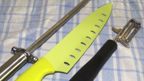 Ein schneidendes Küchengerät, ein Messer, liegt auf einem karierten Küchenhandtuch, umgeben von einem Wetzstahl und einem Sparschäler.
