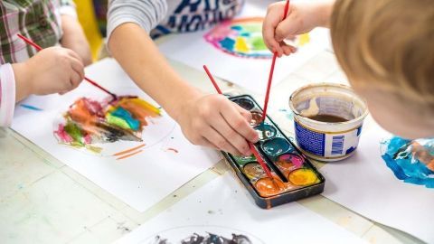 Hände von Kindern malen mit Wasserfarben auf Papier, während eine Farbpalette und ein Wasserglas in der Nähe stehen.