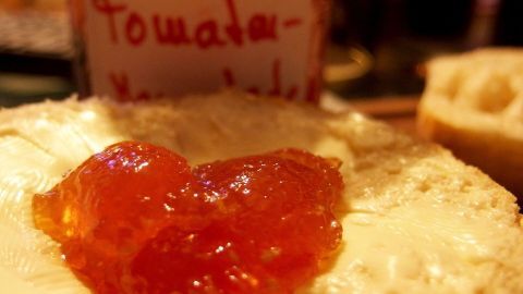 Auf einem Brötchen liegt eine Portion Tomatenmarmelade, daneben steht ein Glas mit beschriftetem Etikett in warmem Licht.